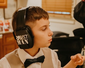 May include: A young person wearing a white shirt and a brown vest with a black bow tie. They are wearing black noise-canceling headphones with the words "Best Man" printed on them.