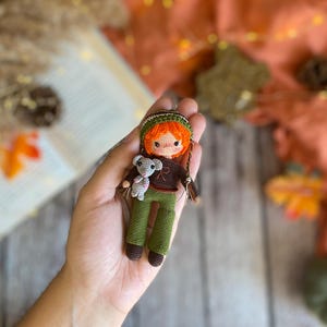 Könnte beinhalten: Eine handgefertigte Häkelpuppe mit orangefarbenem Haar, einem grünen Hut und einer grünen Hose, die einen kleinen grauen Teddybären hält. Die Puppe hat ein braunes Hemd und braune Schuhe. Die Puppe wird in einer Hand gehalten.