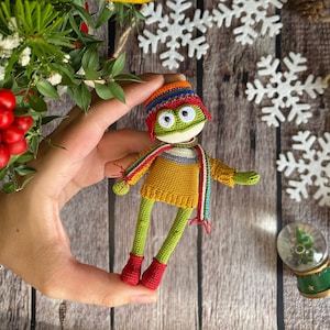 May include: A green crocheted frog wearing a colorful striped hat and scarf, a yellow sweater, and red boots. The frog is being held by a hand against a wooden background with white snowflakes.