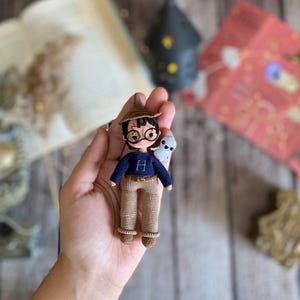 May include: A handmade crochet doll of a character wearing a brown hat, round glasses, and a blue sweater with a white "H" on it. The doll is holding a small white owl. The doll is wearing tan pants and brown shoes.