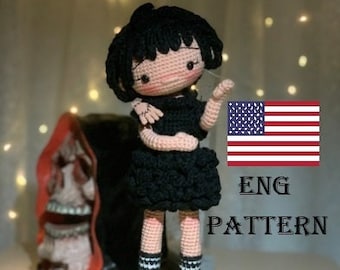 Könnte beinhalten: Häkelpuppenmuster auf Englisch. Die Puppe trägt ein schwarzes Kleid und schwarz-weiß gestreifte Socken. Die Puppe hat schwarze Haare und ein rosafarbenes Gesicht. Die Puppe steht vor einer rot-weiß-blauen amerikanischen Flagge.