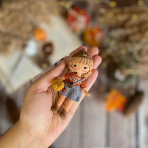 May include: A handmade crochet doll with a straw hat, red shirt, blue shorts, and brown boots. The doll is holding a yellow bag. The doll has blonde hair and is wearing a brown hat. The doll is a miniature version of a scarecrow.