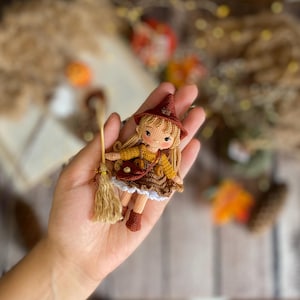 May include: A handmade crochet doll of a witch, featuring a brown pointed hat, blonde hair, and a brown and yellow dress. The doll is holding a small broom. The doll has a small purse and brown boots. The doll is a charming, whimsical decoration.