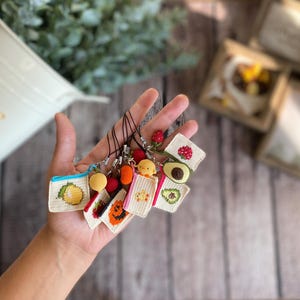 以下が含まれることがあります： レモン、イチゴ、カボチャ、ヒヨコ、アボカドなどの様々なデザインが施された、小さなかぎ針編みのポーチとチャームのコレクション。各アイテムは、銀色の金属製クラスプが付いた黒いコードループに取り付けられています。