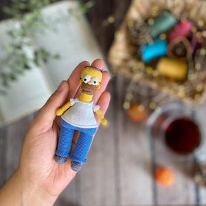 May include: A crocheted yellow character with blue trousers and a white shirt. The character has a brown moustache and glasses. The character is being held in a person's hand.
