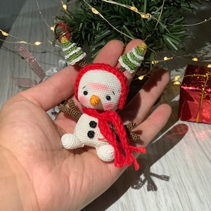 May include: A small crocheted snowman ornament with a red hat, scarf, and green tree-shaped antlers. The snowman has black eyes, a carrot nose, and black buttons.
