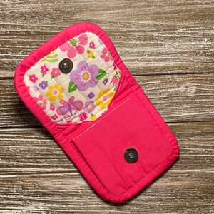 May include: A pink fabric pouch with a floral print and a flap closure. The pouch has a small pocket on the front and a button closure.
