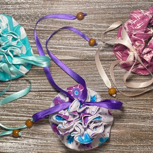 May include: Three fabric drawstring pouches with different patterns and colors. The pouches are tied with satin ribbons and have wooden beads on the ends. The pouches are on a wooden surface.