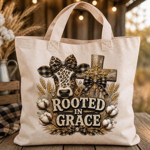 PNG File Transparent Background A beige canvas tote bag featuring a graphic print of a cow with a leopard print pattern and a plaid bow. The design includes a wooden cross, wheat stalks, cotton bolls, and the text ROOTED IN GRACE with additional plaid bows. Digital sublimation design.