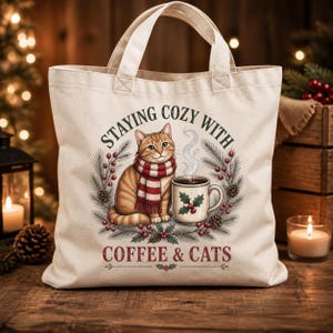 May include: A cream-colored canvas tote bag featuring a graphic of an orange tabby cat wearing a red and white striped scarf next to a steaming mug of coffee. The design includes pine branches, red berries, and the text STAYING COZY WITH COFFEE & CATS.