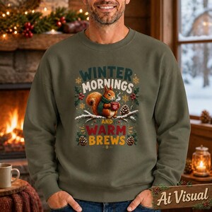 May include: An olive green crewneck sweatshirt featuring a graphic of a squirrel wearing a green scarf and holding a red mug. The text reads Winter Mornings and Warm Brews, surrounded by illustrations of pinecones, holly berries, and snowflakes.