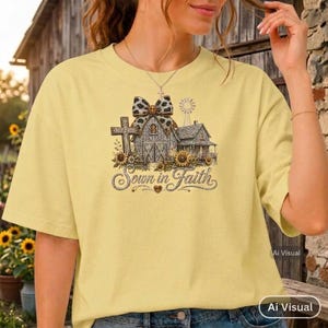 May include: A pale yellow t-shirt featuring a graphic print of a barn, a cross, and a windmill decorated with a leopard print bow and sunflowers. The text "Sown in Faith" is printed in a cursive script below the barn and cross illustration.