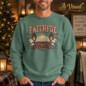 May include: A sage green crewneck sweatshirt featuring a graphic print of an open book, two lanterns, and cotton sprigs. The text reads FAITHFUL in a patterned block font and Heart in a red script font, centered on the chest of the garment.