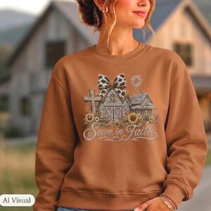 May include: A brown crewneck sweatshirt featuring a graphic print of a barn, a cross, and a windmill, all topped with a large leopard-print bow. The design includes sunflowers and the cursive text Sown in Faith written in a light, metallic-toned script.
