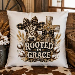 May include: A white square throw pillow featuring a central graphic of a cow with a leopard print head and black and tan plaid ears. The cow wears a matching bow. Beside it is a wooden cross with a plaid bow, wheat stalks, and cotton bolls.