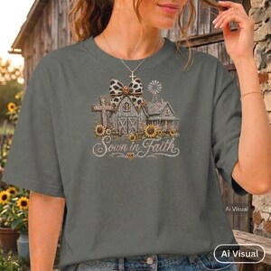May include: A charcoal gray short-sleeve t-shirt featuring a graphic print of a farmhouse, a cross, and a windmill. The design includes a leopard print bow, sunflowers, and the text Sown in Faith written in a cursive script font across the center.