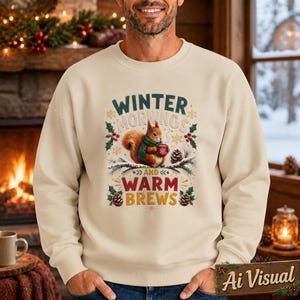 May include: A beige crewneck sweatshirt featuring a graphic of a squirrel wearing a green scarf and holding a red mug. The text reads "WINTER MORNINGS AND WARM BREWS" in green and red lettering, surrounded by pinecones, holly, and snowflake accents.