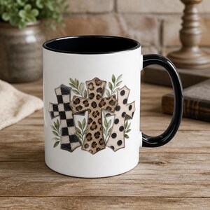 Pu&ograve; includere: Una tazza in ceramica bianca con interno e manico neri. L'esterno presenta una grafica di tre croci decorate con motivi a scacchi bianchi e neri, stampa leopardata marrone e pois beige, accentuati da rametti di foglie verdi.