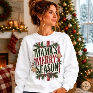 May include: A white long-sleeve sweatshirt featuring a central graphic with the text MAMA'S MERRY SEASON in red and green lettering. The design includes illustrations of holly leaves, red berries, and gingerbread stars on a striped background.