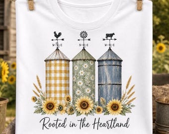 Puede incluir: Una camiseta blanca con un gráfico de tres silos coronados por veletas. Los silos tienen patrones de cuadros amarillos, flores blancas y madera azul. Girasoles y tallos de trigo rodean la base, con el texto Rooted in the Heartland.