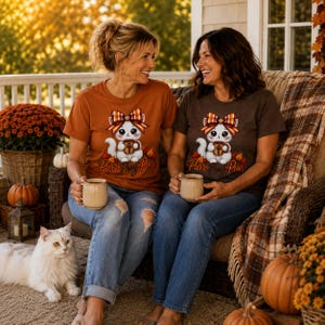 K&ouml;nnte beinhalten: Zwei kurz&auml;rmelige T-Shirts, eines in gebranntem Orange und eines in Dunkelbraun, mit einer Grafik eines wei&szlig;en K&auml;tzchens, das eine karierte Schleife tr&auml;gt und eine Tasse h&auml;lt, sowie dem Schriftzug Purr-Pray-Pour in kursiver Schrift.