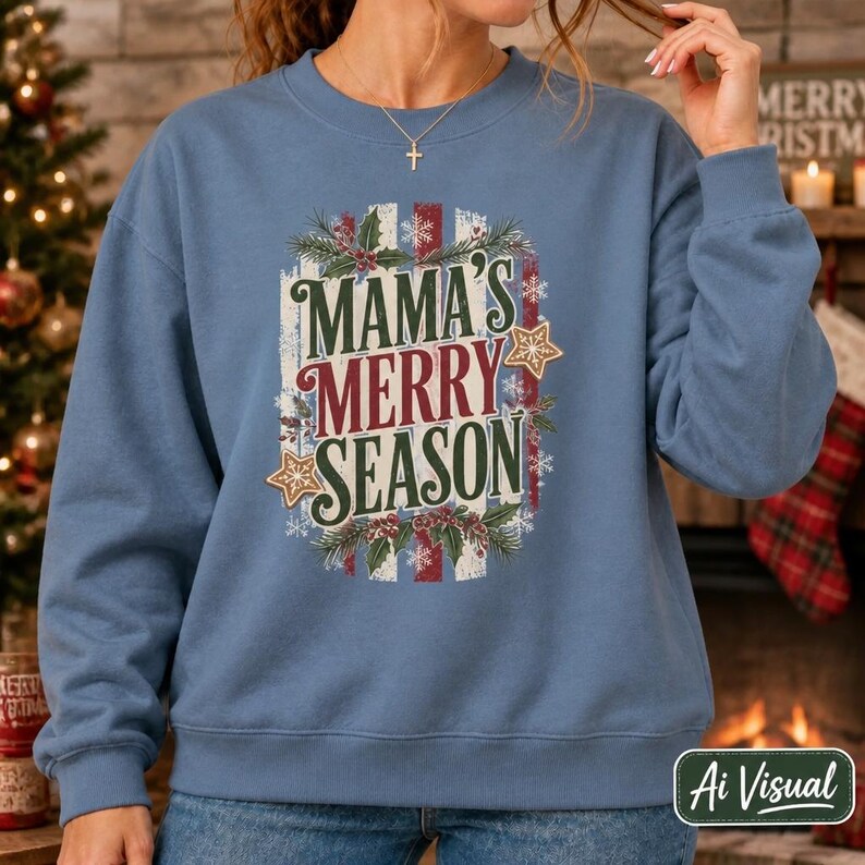 May include: A dusty blue crewneck sweatshirt featuring a central graphic with the text MAMA'S MERRY SEASON in dark green and red lettering. The design is surrounded by illustrations of holly leaves, red berries, snowflakes, and gold stars.