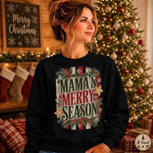 May include: A black long-sleeve sweatshirt featuring a central graphic with the text MAMA'S MERRY SEASON in green and red lettering. The design includes illustrations of pine branches, red holly berries, white snowflakes, and two gold stars.