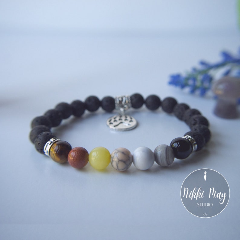 Puede incluir: Pulsera de roca de lava negra con un dije de pata de plata y cuentas de piedras preciosas de colores. Las cuentas son marrones, rojas, amarillas, blancas, grises y negras.