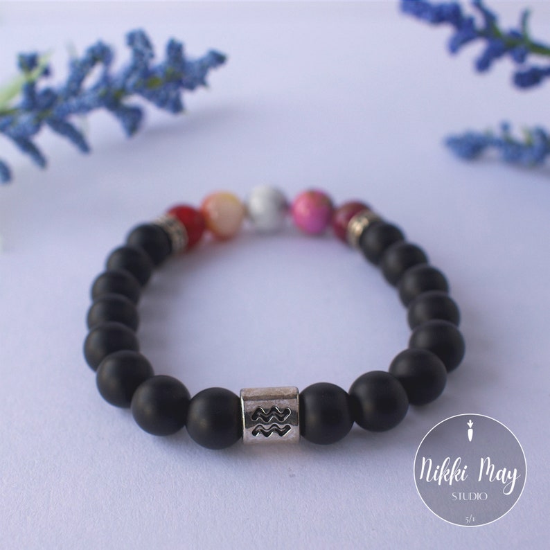 May include: Black beaded bracelet with a silver charm featuring the Aquarius zodiac symbol. The bracelet also has colorful beads in red, pink, white, and orange.