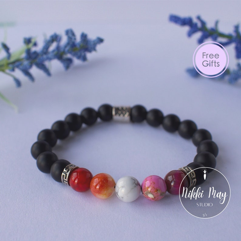 May include: Black beaded bracelet with a silver charm and four colorful gemstone beads. The bracelet is on a white background with a lavender sprig and a pink circle with the text "Free Gifts".