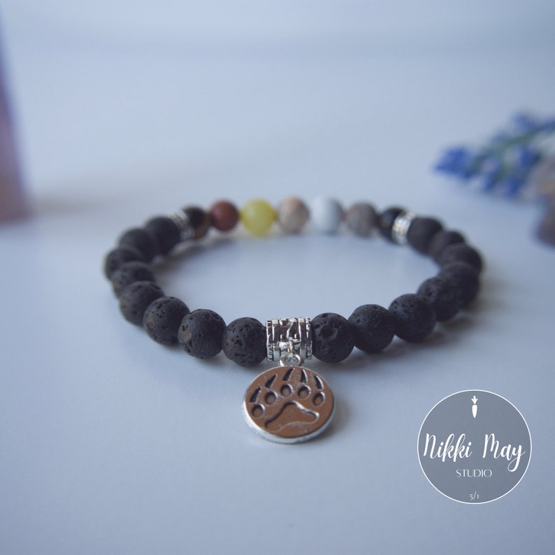 Puede incluir: Pulsera de cuentas de roca volc&aacute;nica negra con un dije de plata en forma de pata de oso. La pulsera tiene una variedad de peque&ntilde;as cuentas de colores intercaladas.
