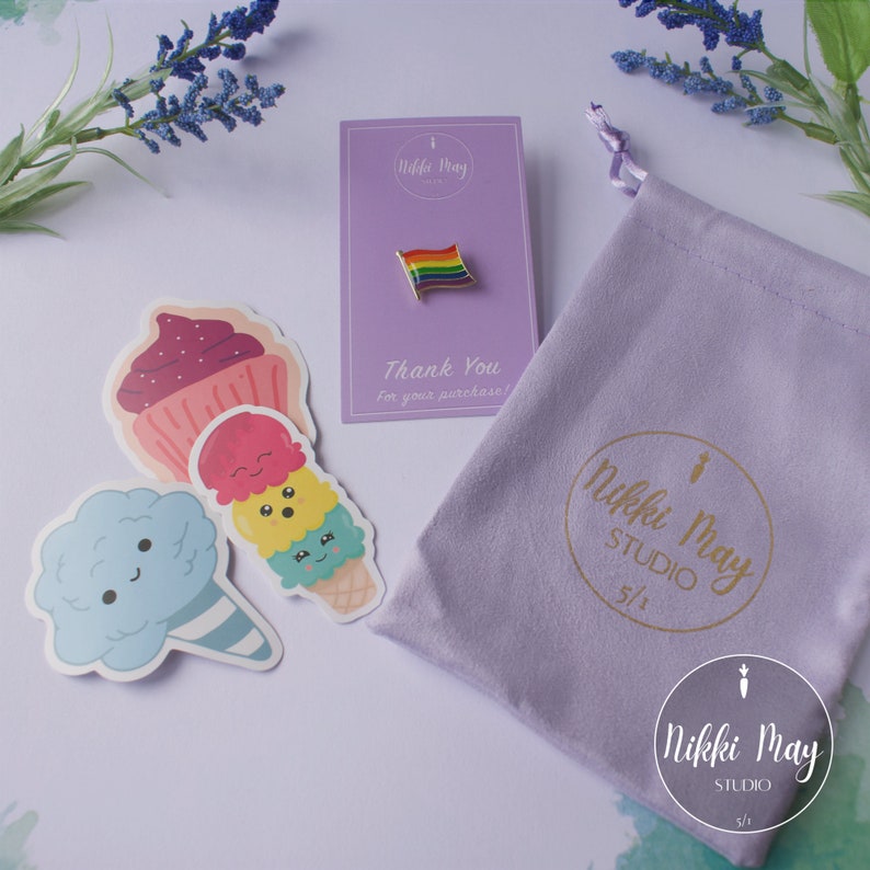 Puede incluir: Una bolsa de terciopelo morado con un logotipo dorado que dice "Nikki May Studio 5/1". La bolsa est&aacute; al lado de una tarjeta con un pin de bandera arco&iacute;ris y el texto "Nikki May Studio" y "&iexcl;Gracias por su compra!" Hay tres pegatinas con im&aacute;genes de dibujos animados de una magdalena, un cono de helado y algod&oacute;n de az&uacute;car.