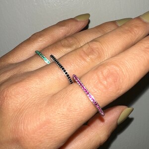 May include: Three thin silver rings with a single row of small gemstones. The rings are on a person's finger. The gemstones are green, black, and pink.
