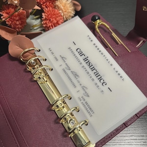 May include: A burgundy binder with gold fittings holds a card that reads "car insurance." The card is translucent with black text. A gold hairpin with a black bead sits in the binder's pocket. Floral arrangement in the background.