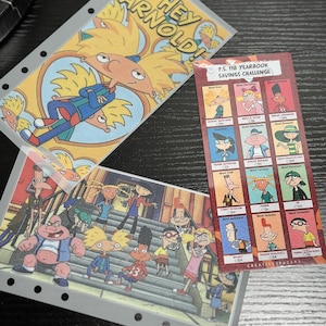 Puede incluir: Tres láminas de plástico transparente con imágenes de dibujos animados de la serie "¡Oye Arnold!". Una lámina presenta a Arnold con el texto "Hey Arnold!". Otra muestra un grupo de personajes. Una tercera tarjeta es un "Yearbook Savings Challenge".