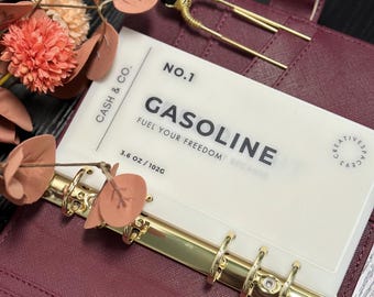 May include: A burgundy binder with gold hardware holds a translucent card labeled "GASOLINE" with the text "FUEL YOUR FREEDOM". A gold hairpin with a black bead sits on the binder. The card is labeled "102g".