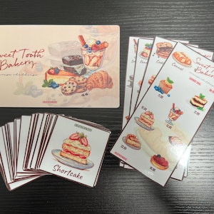 May include: A selection of illustrated cards and a larger sign featuring desserts. The sign reads "Sweet Tooth Bakery" with images of pastries, cakes, and a parfait. Individual cards display shortcake and other desserts.