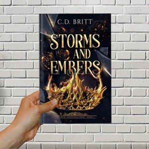 May include: Book cover with a gold crown and flames on a black background. The title is "Storms and Embers" with the author's name "C.D. Britt" above the title. The text "Reign of Goddesses Series - Book 3" is at the bottom of the cover.
