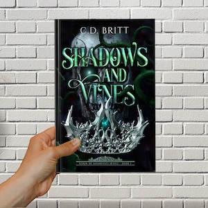 May include: Book cover with a skull and vines, a silver crown with a green gem, and the title "Shadows and Vines" in green text. The book is part of the "Reign of Goddesses Series - Book 1".