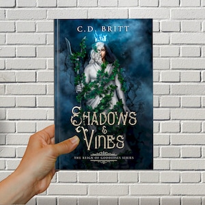 May include: Book cover with a blue and green background featuring a figure with a white face and a crown of vines. The title "Shadows & Vines" is written in gold lettering with the subtitle "The Reign of Goddesses Series".