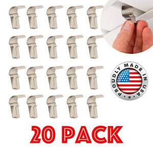 May include: A pack of 20 silver-colored plastic hooks, designed for hanging items from a ceiling. The hooks are shaped like a 'J' and have a small lip on the top to help secure them in place. The hooks are made in the USA and are labeled as a 20 pack.