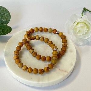 May include: Two brown beaded bracelets on a white marble surface. The bracelets are made of small, round beads and are stacked on top of each other.