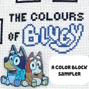 May include: Cross-stitch sampler with the text "THE COLOURS OF BLUEY" and a blue cartoon dog design. Two cartoon dog pins are at the bottom. The text "A COLOR BLOCK SAMPLER" is in a white oval.