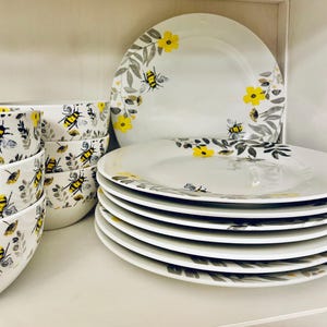 Può includere: Un set di stoviglie in ceramica bianca con un motivo di api gialle, fiori giallo brillante e rami con foglie grigie. La collezione comprende una pila di piatti piani rotondi e una pila verticale di ciotole abbinate con lo stesso motivo.