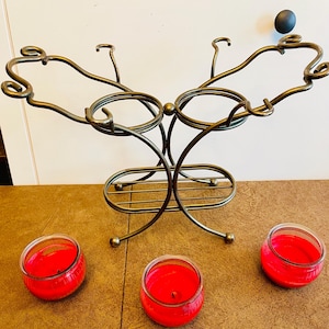 May include: A bronze-toned metal stand featuring two circular holders, curved decorative arms, and a lower shelf, accompanied by three small glass jars filled with bright red wax, arranged in a semi-circular formation in front of the metal structure.