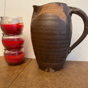Puede incluir: Una jarra de cerámica alta de color marrón oscuro con una superficie texturizada y estriada y un asa grande, junto a una pila vertical de tres frascos de vidrio transparente llenos de cera roja brillante, con nervaduras verticales.