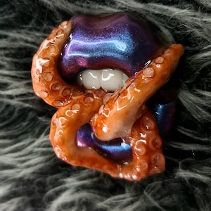 May include: A close-up of a purple and blue iridescent lip sculpture with an octopus tentacle wrapped around it.