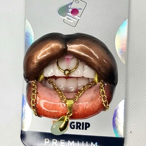 Puede incluir: Un agarre para tel&eacute;fono con una escultura 3D realista de labios de color marr&oacute;n oscuro con dientes blancos, un anillo de septum dorado y un collar de cadena dorada con un peque&ntilde;o colgante verde p&aacute;lido. El empaque incluye el texto GRIP y PREMIUM.
