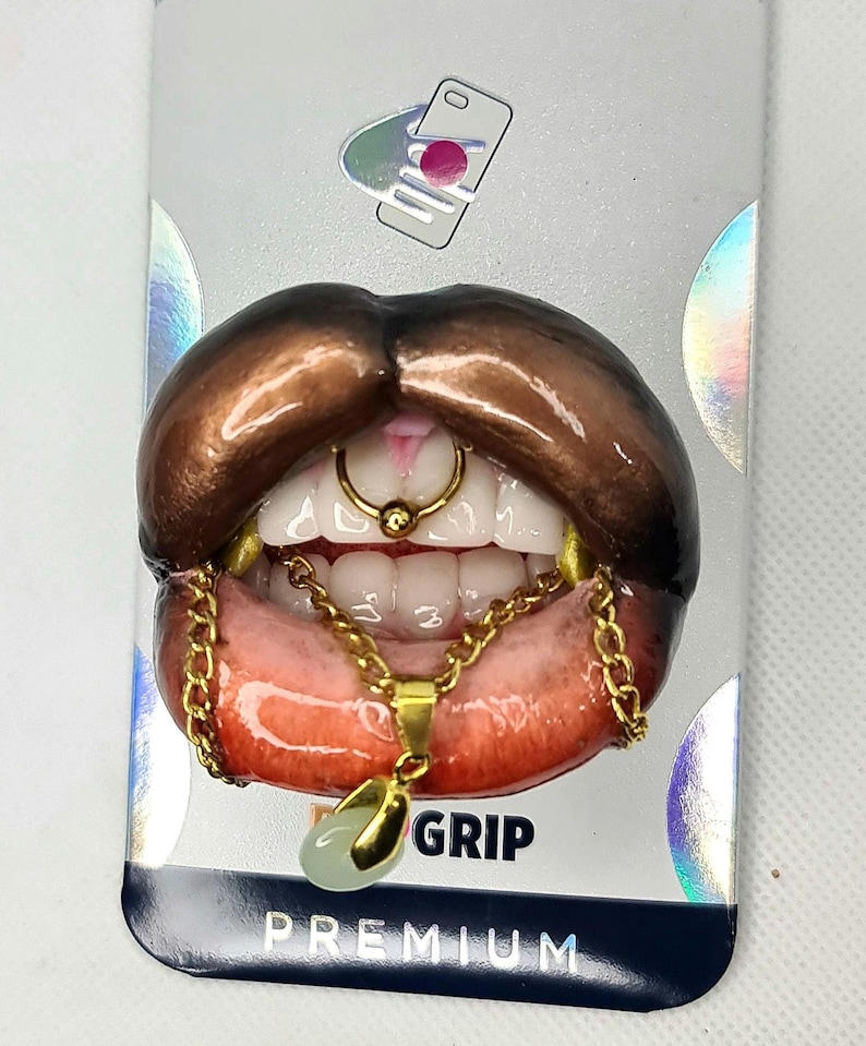 Puede incluir: Un agarre para tel&eacute;fono con una escultura 3D realista de labios de color marr&oacute;n oscuro con dientes blancos, un anillo de septum dorado y un collar de cadena dorada con un peque&ntilde;o colgante verde p&aacute;lido. El empaque incluye el texto GRIP y PREMIUM.