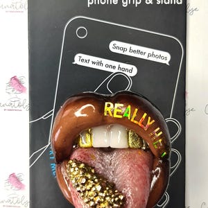 Puede incluir: Un agarre para teléfono y un soporte con un diseño de labios marrones y rosados con dientes de oro y una lengua cubierta de purpurina dorada. El texto "REALLY HE" está escrito en los labios.